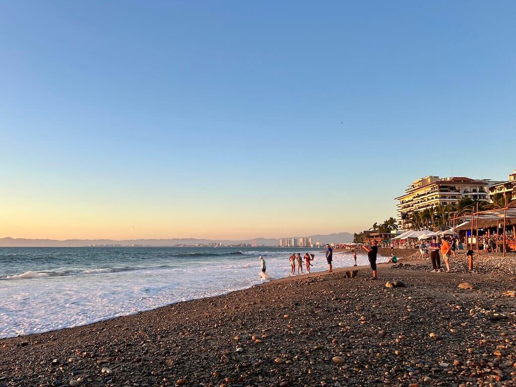 beachgoers congregate on playa camarones in Puerto Vallarta to capture photos of the sunset. looking north along the beach with the ocean lapping the shore on the left side of the image