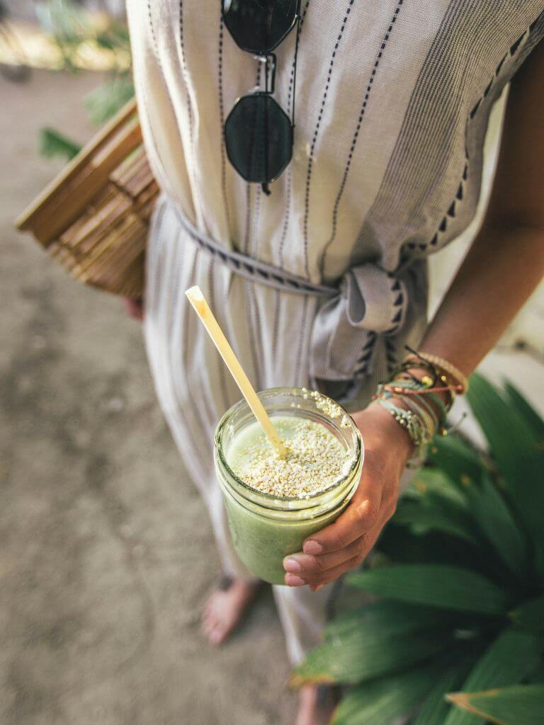 a woman in a gauzy white dress with black embroidery holds a green matcha drink in a mason jar.